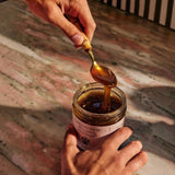 Hands cradle the Flamingo Estate California Mountain Wildflower Honey jar as a brass spoon drizzles thick golden honey back inside.