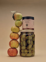 A Flamingo Estate California Mountain Wildflower Honey jar balances atop a jar of green olives next to a totem-stack of apples.