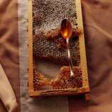 A wooden bee-frame holds raw honeycomb on jute, a brass spoon catching golden Flamingo Estate California wildflower honey.