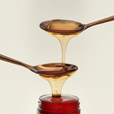 Two spoons cascade Flamingo Estate California wildflower honey, pale gold dripping into darker amber and into the open jar below.