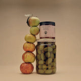A Flamingo Estate California Mountain Wildflower Honey jar balances atop a jar of green olives next to a totem-stack of apples.