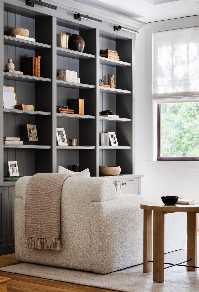 Charcoal library shelving styled with books and ceramics behind a boucle armchair and oak side table