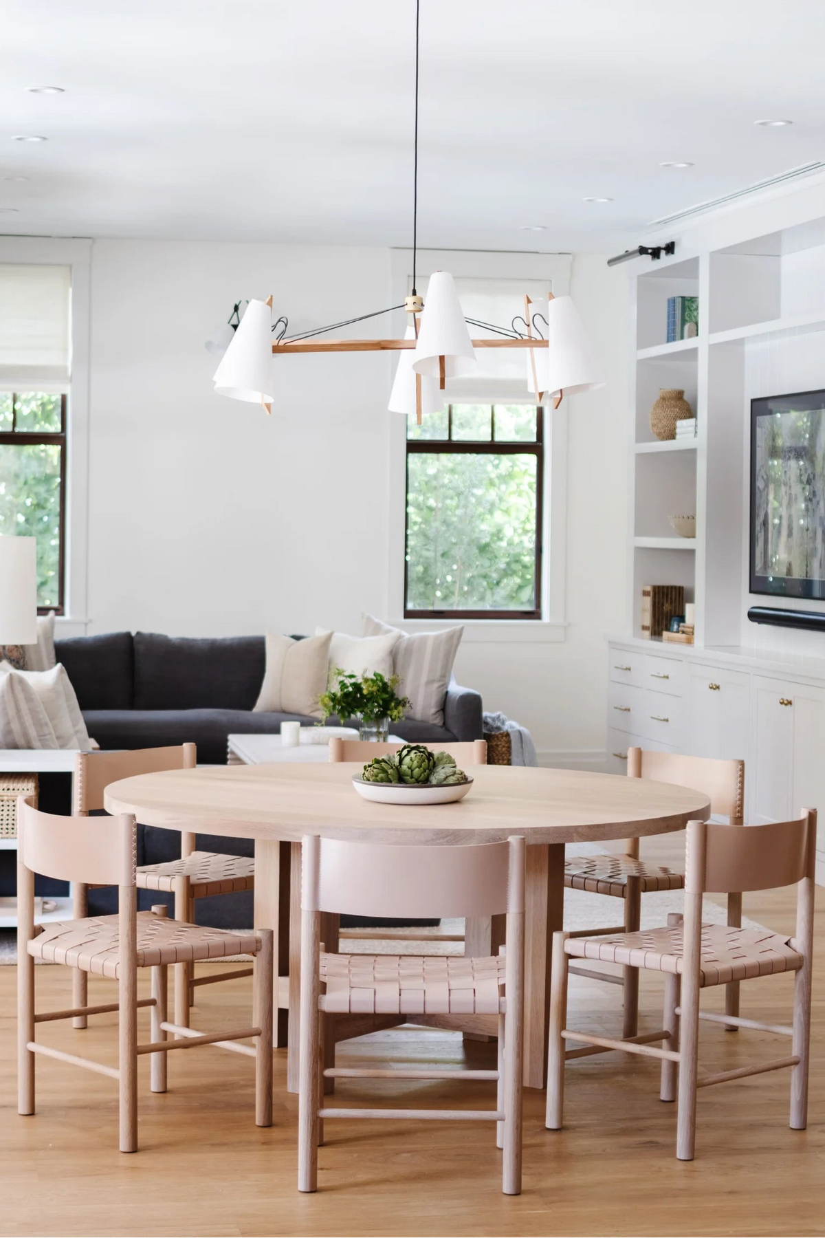A round light wood dining table with six matching wooden chairs featuring woven seat slats, shown in a bright, modern living room setting.