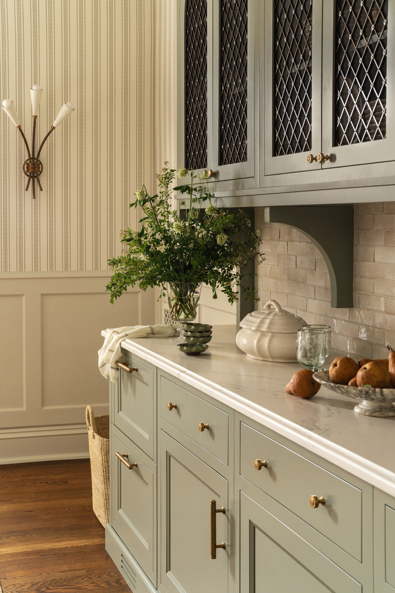 Butler's pantry detail with sage green cabinetry, a brass sconce, and greenery on the marble counter