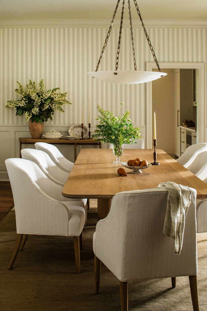 Dining room with an oak table, slipcovered chairs, and an alabaster pendant over striped wallpaper