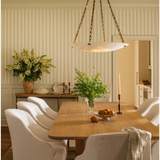 A dining room scene with a light wood rectangular table surrounded by white upholstered chairs, a hanging pendant light, striped wallpaper, and floral arrangements in the background.