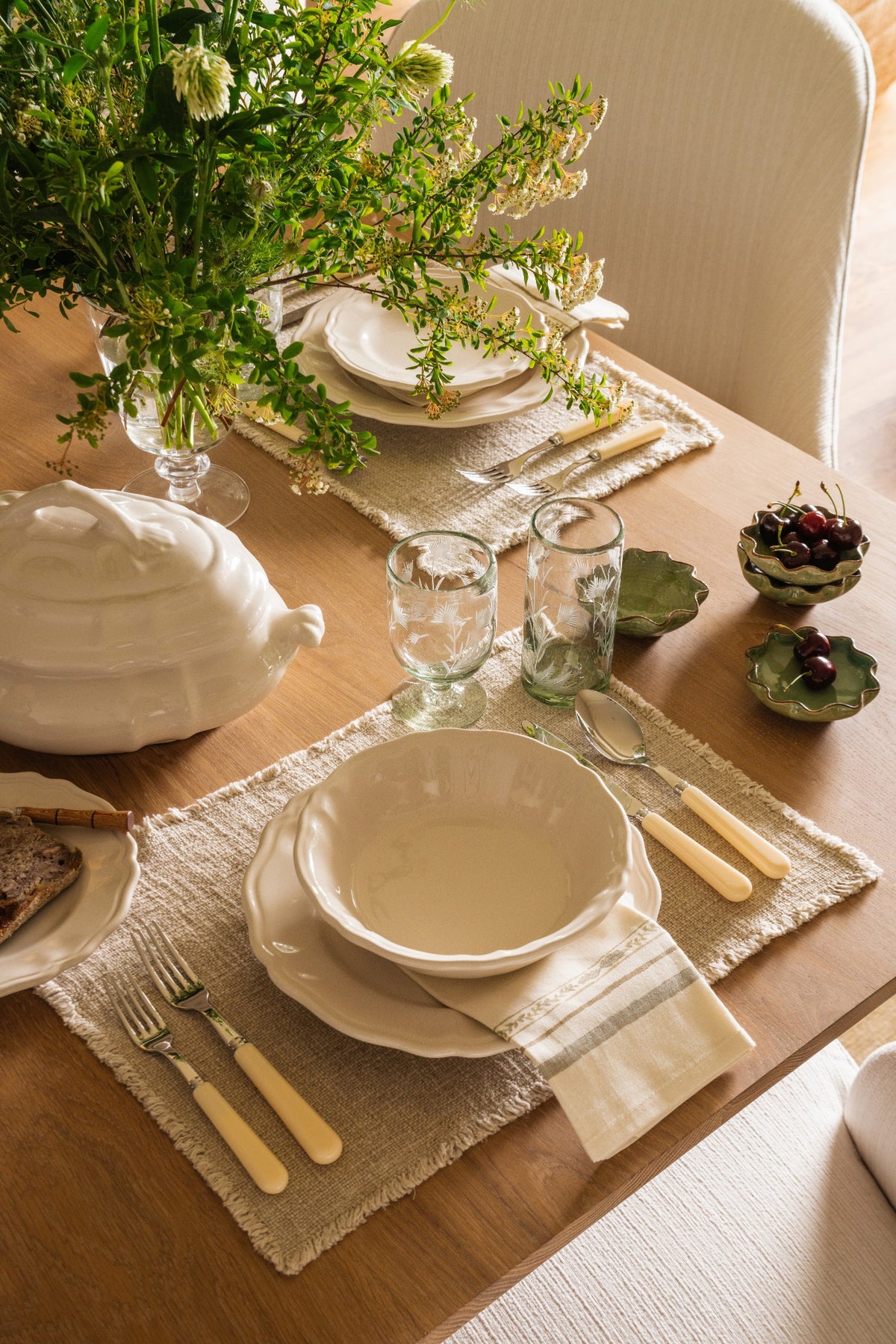 Place setting detail with a cream tureen, gold flatware, linen runner, and greenery on an oak table