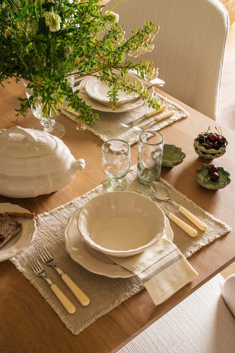 Place setting detail with a cream tureen, gold flatware, linen runner, and greenery on an oak table