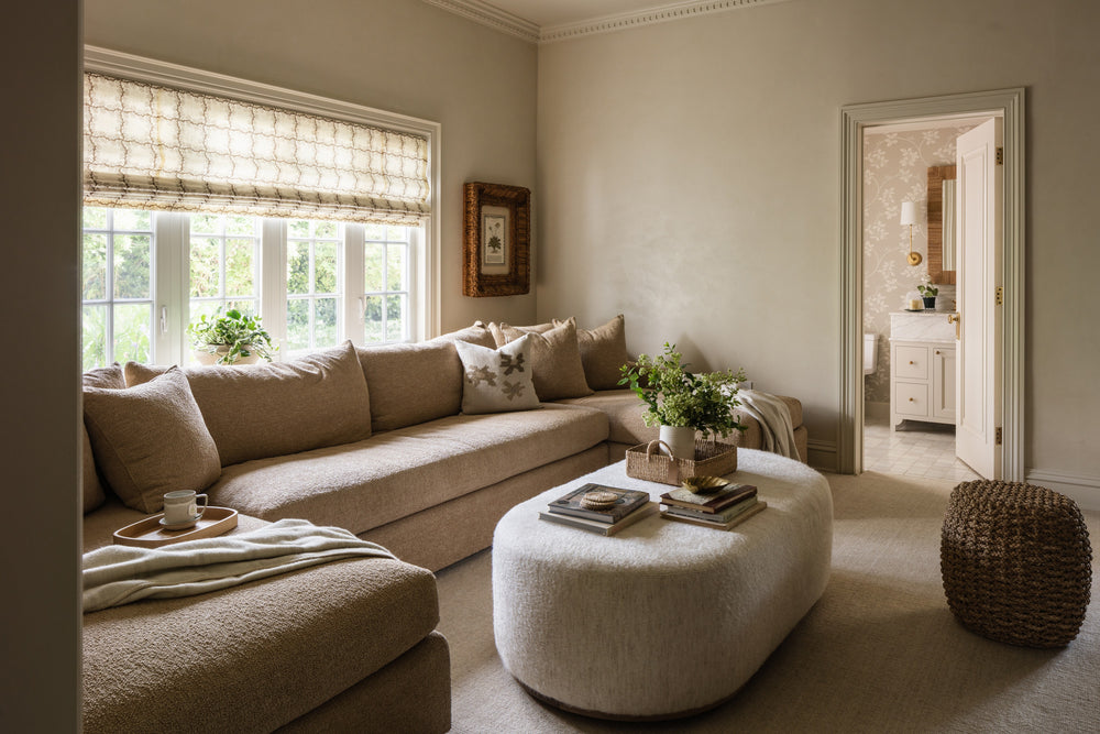 Family room with a linen sectional, oval bouclé ottoman, woven pouf, and patterned roman shade