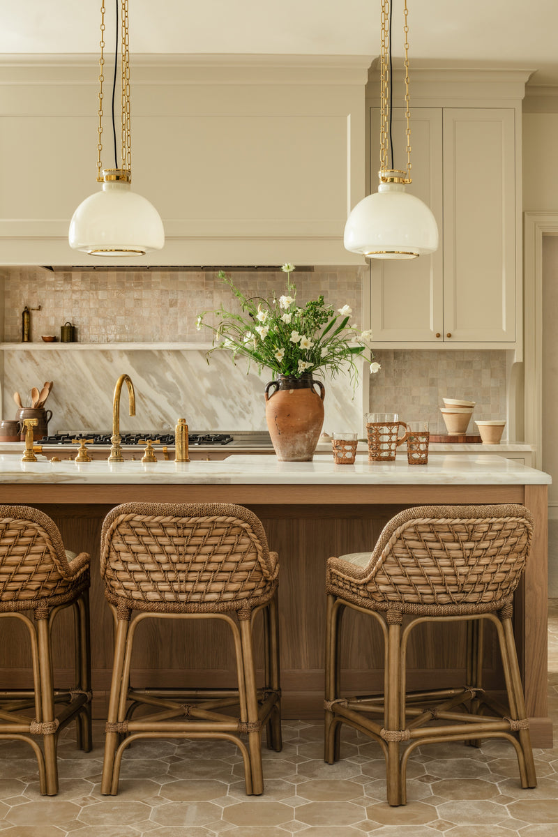 Kitchen island with rattan counter stools, marble backsplash, and brass fixtures