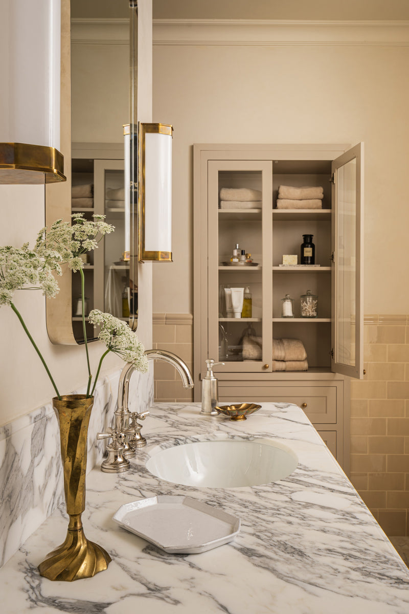 Primary bathroom detail with a marble counter, brass fittings, and a linen-shelved cabinet