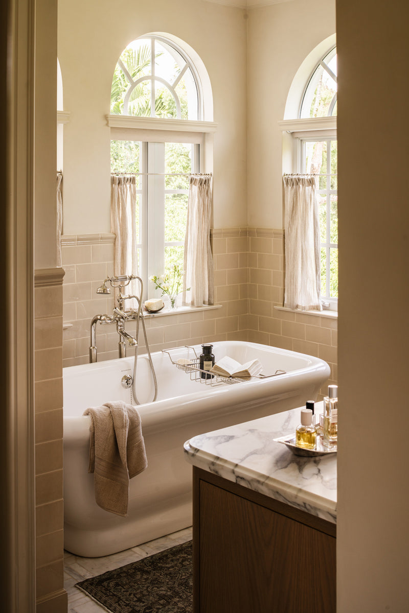 Primary bathroom with a freestanding soaking tub beneath arched windows and an oak vanity