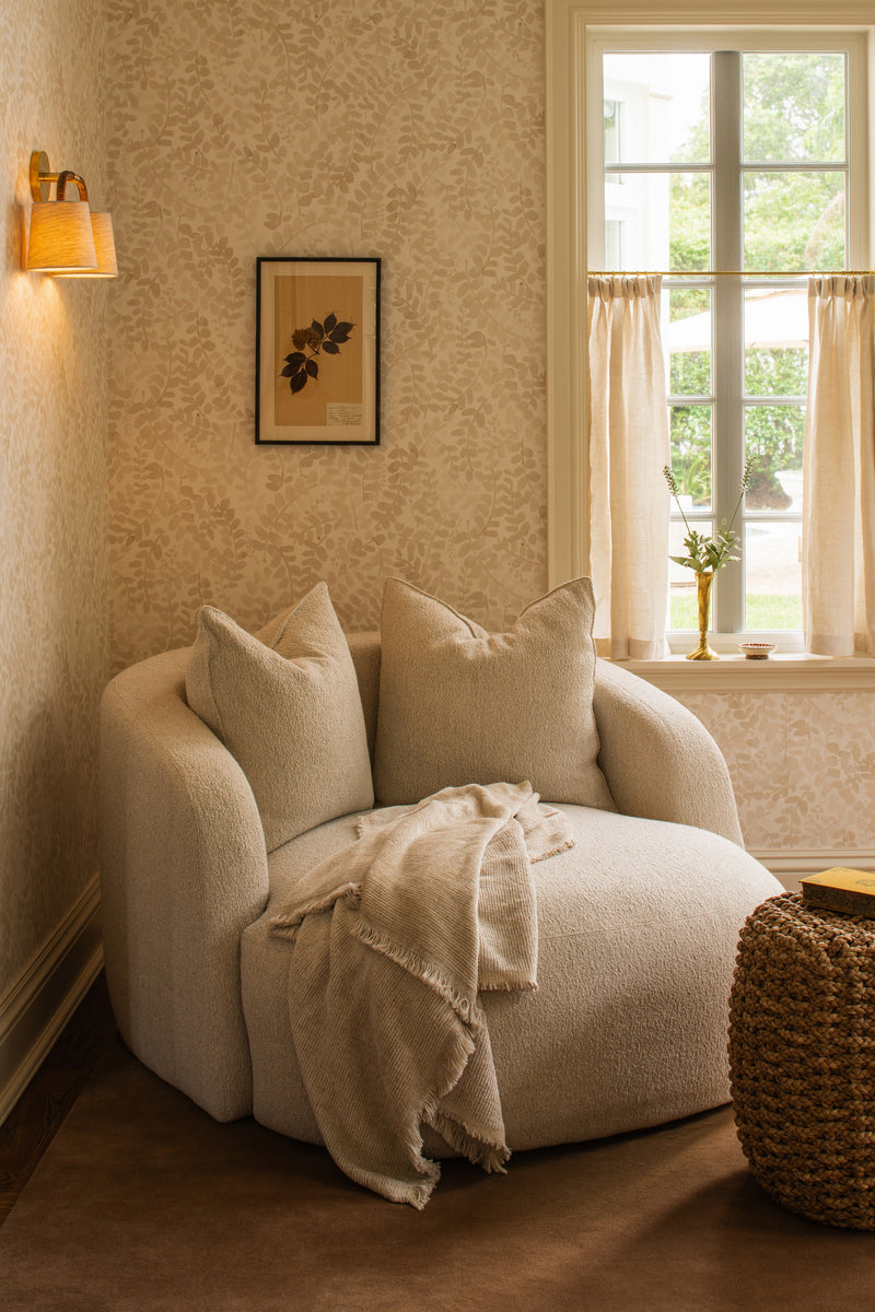 Reading nook with a curved bouclé chair, woven side table, and brass sconce beside an arched window