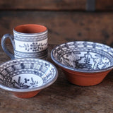 Calimia Home hand-thrown ceramic bowls and mug with grey floral glaze and terracotta base on a rustic wooden table.