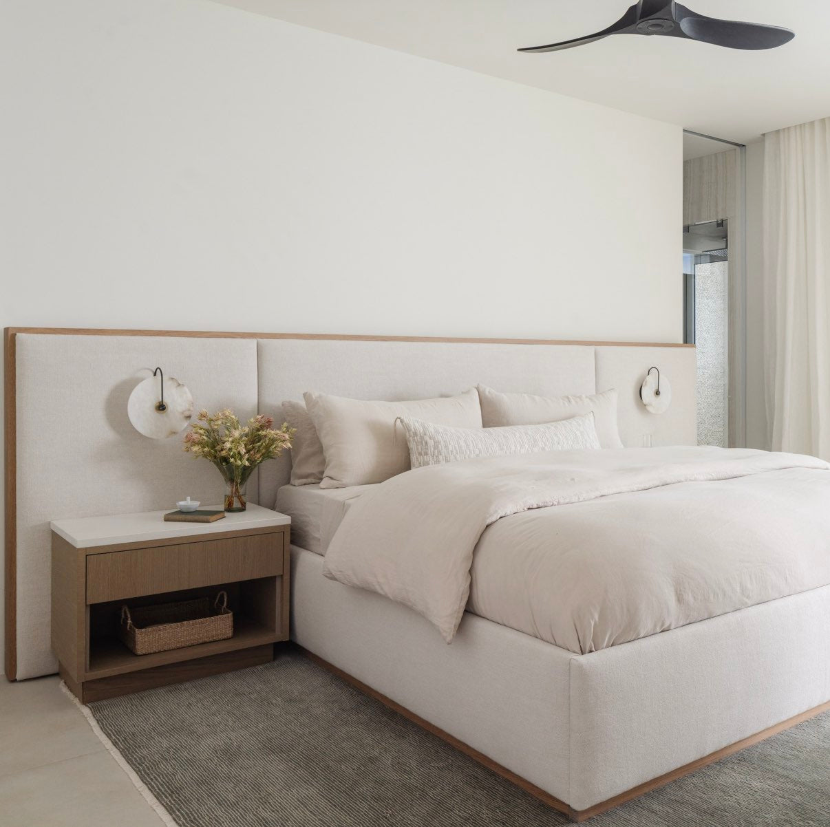 A serene neutral bedroom with a wide upholstered bed, oak nightstand, and alabaster wall sconce.