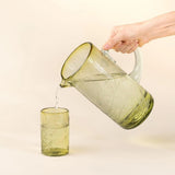 Calimia Home hand-blown olive recycled glass pitcher with etched thistle design pouring water into a matching glass