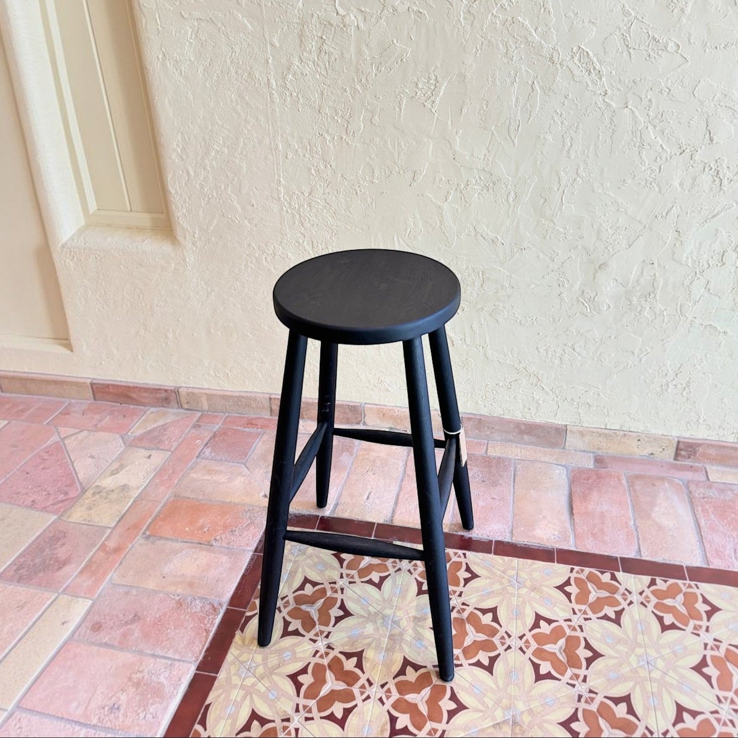 Calimia Home Omah stool in black stained wood with a round seat and tapered legs on a tiled floor.