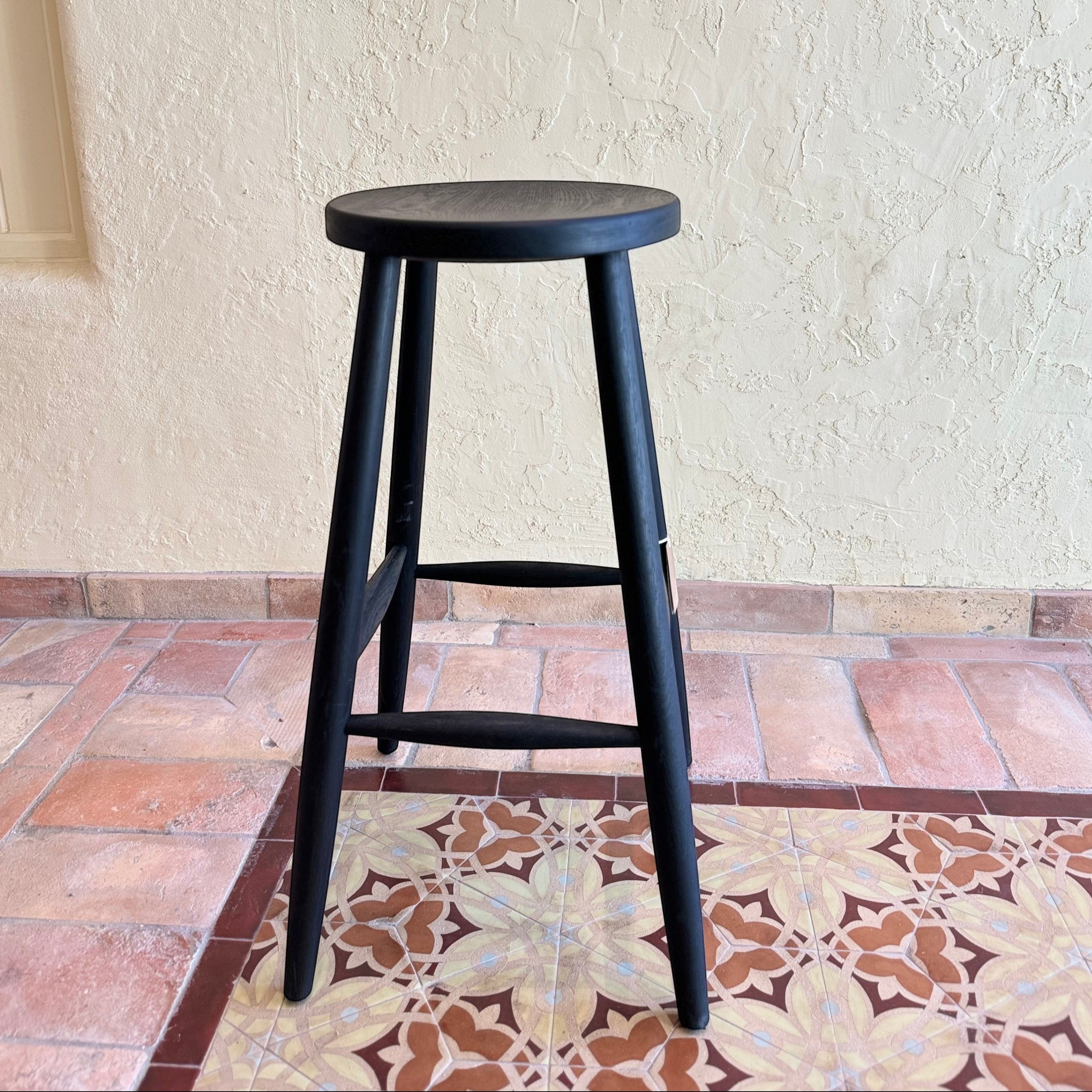Calimia Home Omah stool in black stained wood with a round seat and tapered legs on a patterned tile floor.