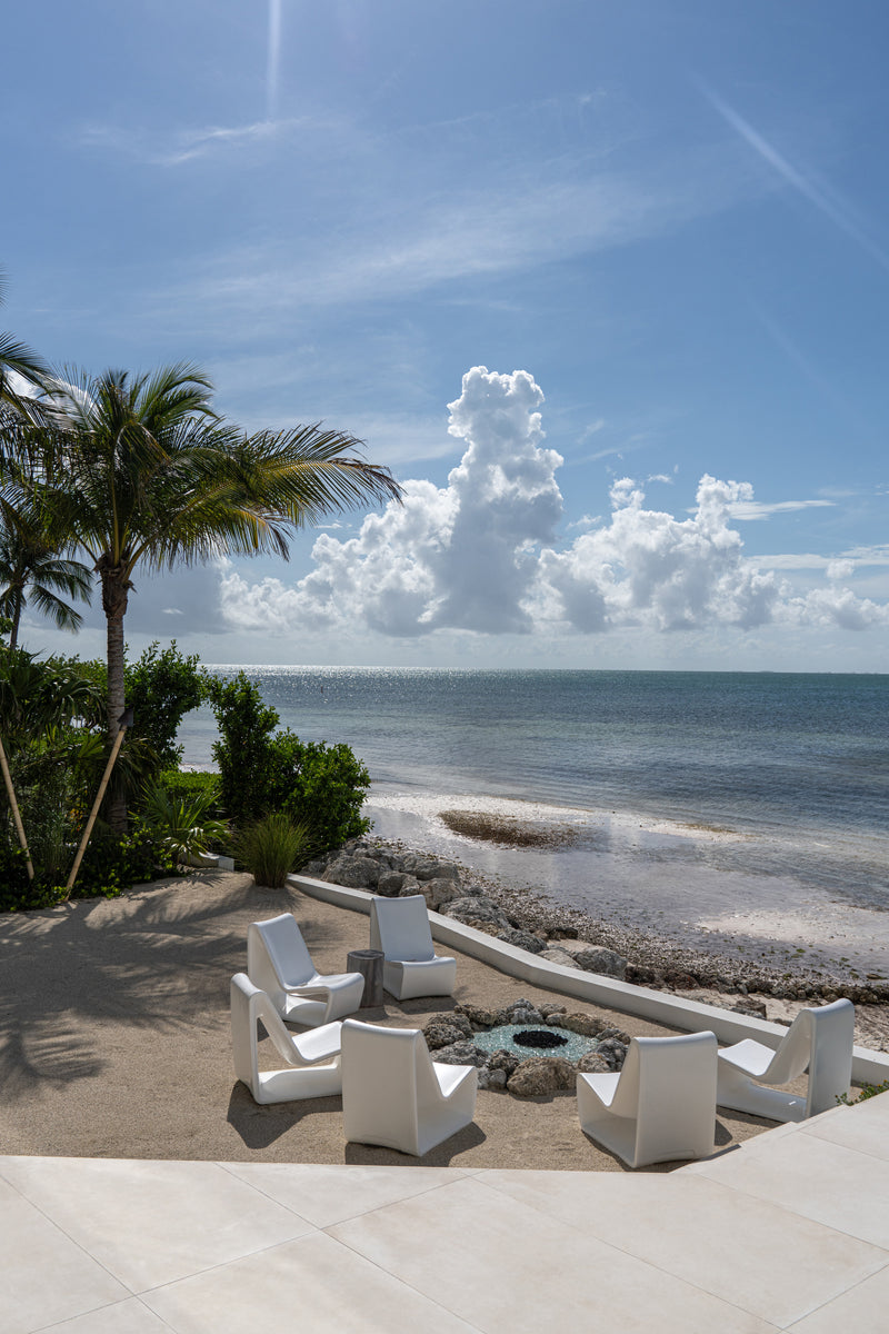 Sandy beachfront firepit lounge with white Adirondack chairs beneath a palm tree
