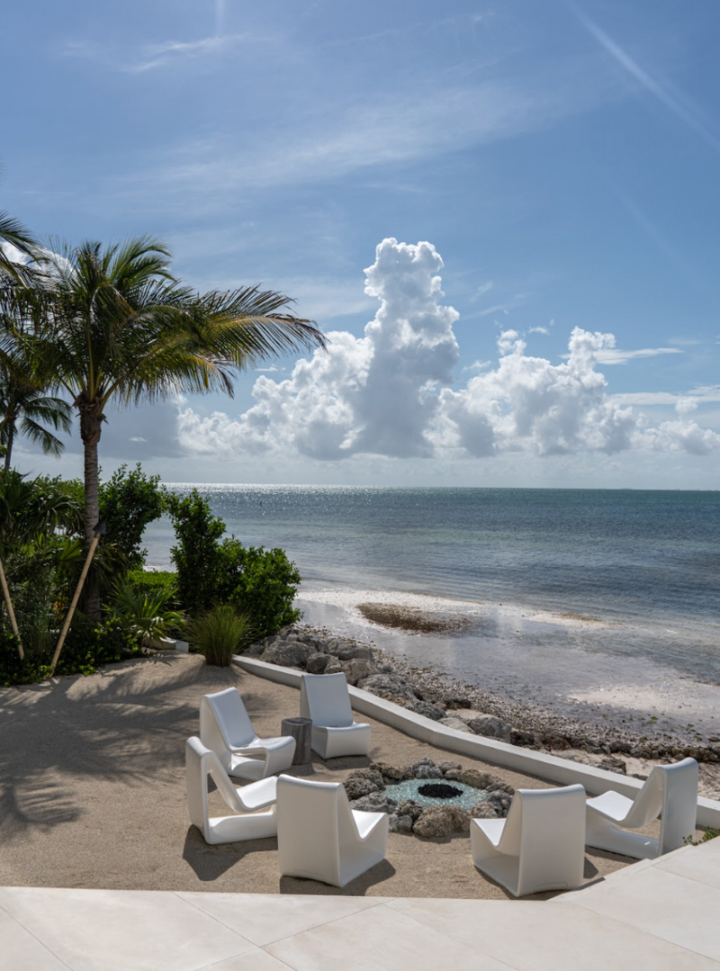 Sandy beachfront firepit lounge with white Adirondack chairs beneath a palm tree