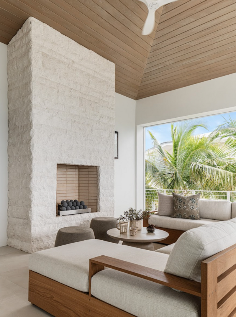 Covered lanai with whitewashed stone fireplace, teak sofas, and palm views designed by Calimia Home
