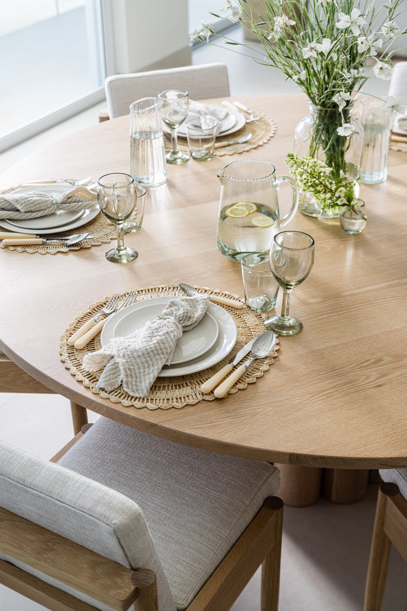 Round oak table set with rattan chargers, fringed napkins, and fresh greenery