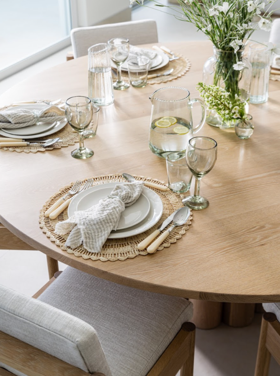 Round oak table set with rattan chargers, fringed napkins, and fresh greenery