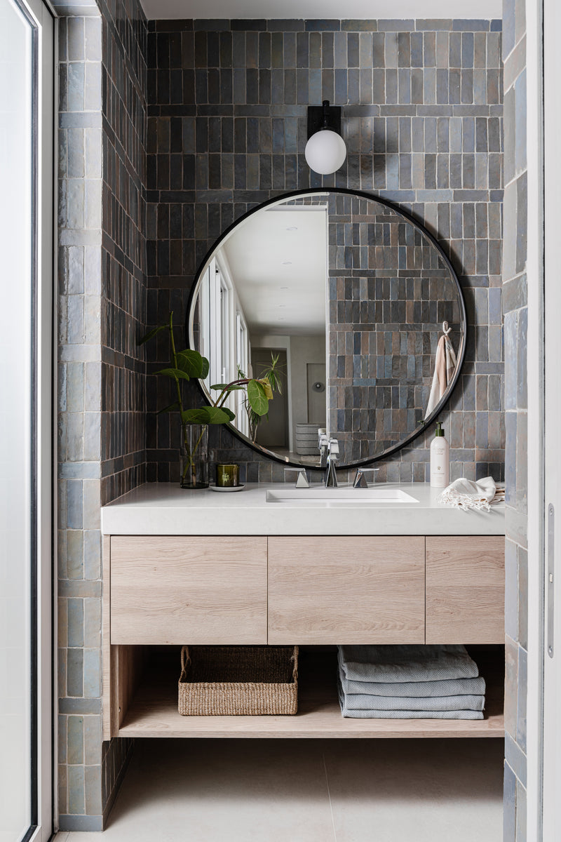 Powder bath with blue-gray zellige tile, round black mirror, and floating white oak vanity