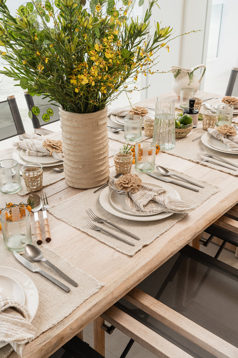 Tablescape detail with woven placemats, ribbed vase of yellow blooms, and mixed glassware