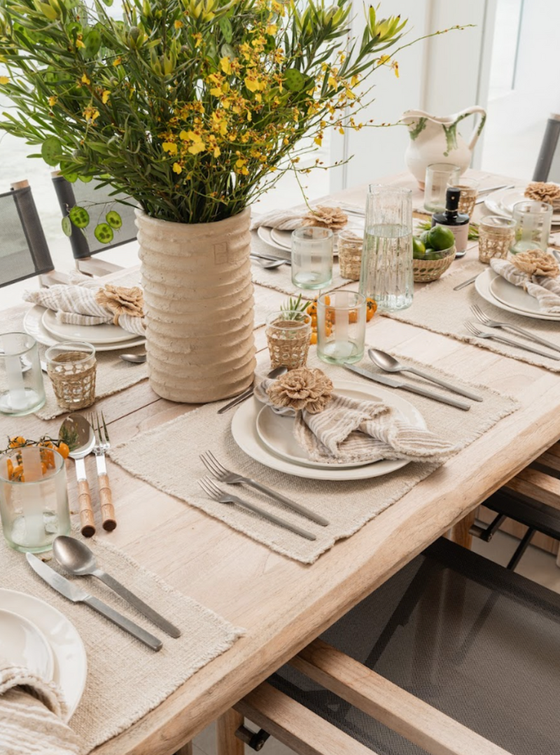 Tablescape detail with woven placemats, ribbed vase of yellow blooms, and mixed glassware