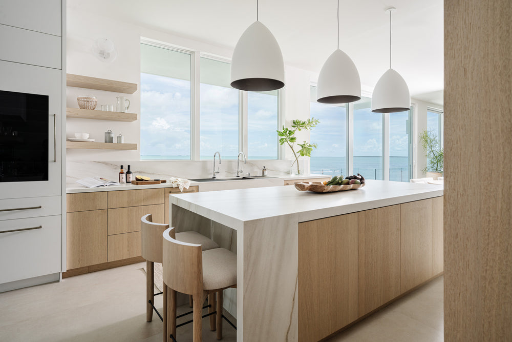 White oak kitchen with waterfall-edge island, dome pendants, and ocean views beyond the windows