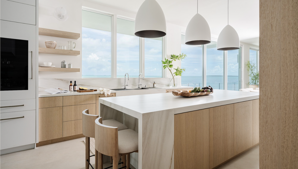 White oak kitchen with waterfall-edge island, dome pendants, and ocean views beyond the windows