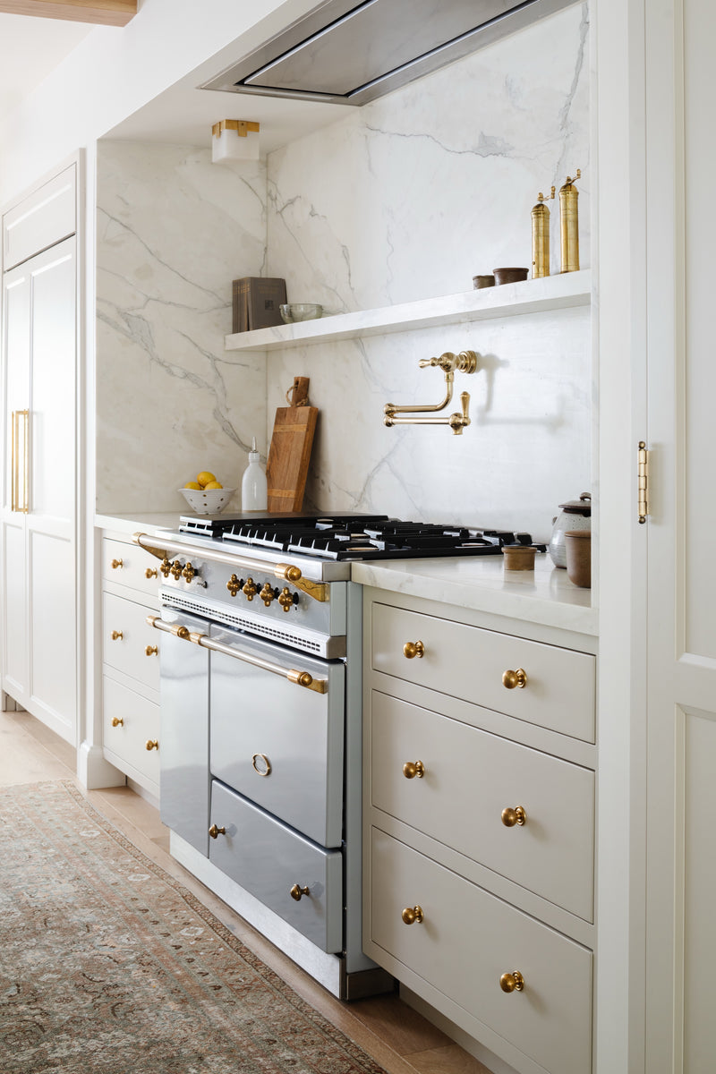 Cream kitchen cabinetry with brass hardware, marble backsplash, and pot filler above the range