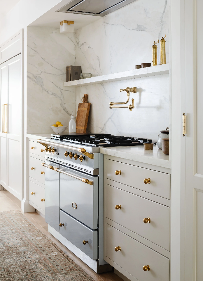 Cream kitchen cabinetry with brass hardware, marble backsplash, and pot filler above the range