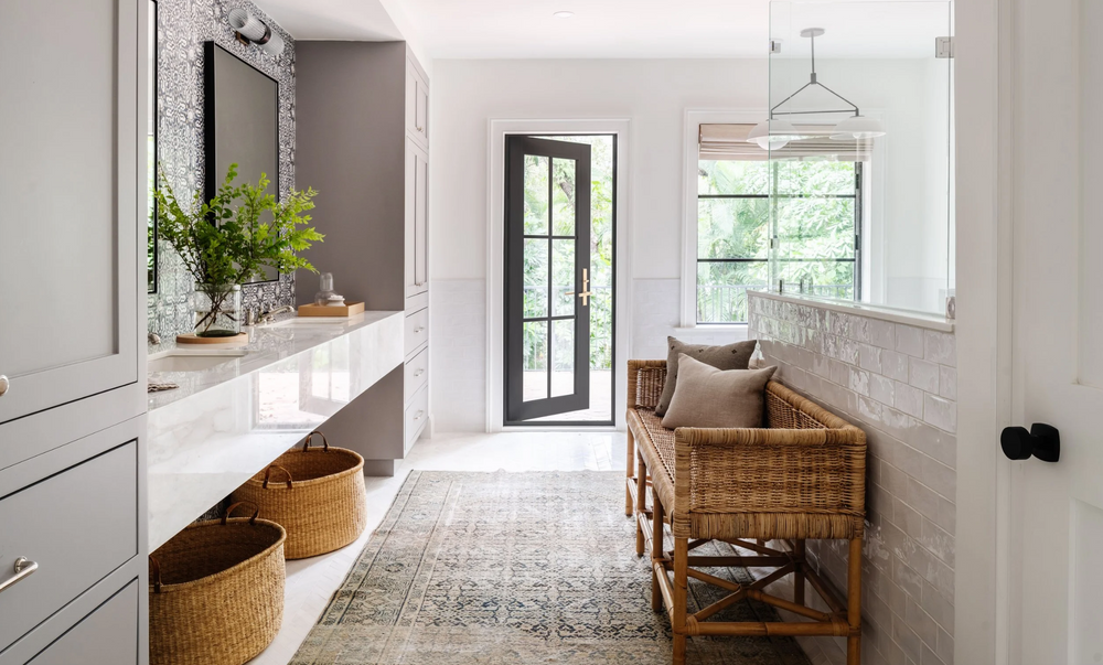 Primary bath with long white vanity, rattan bench, and vintage runner designed by Calimia Home