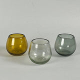 Calimia Home Roli Poli stemless glasses in amber, clear, and grey hand-blown recycled glass with a rounded shape.