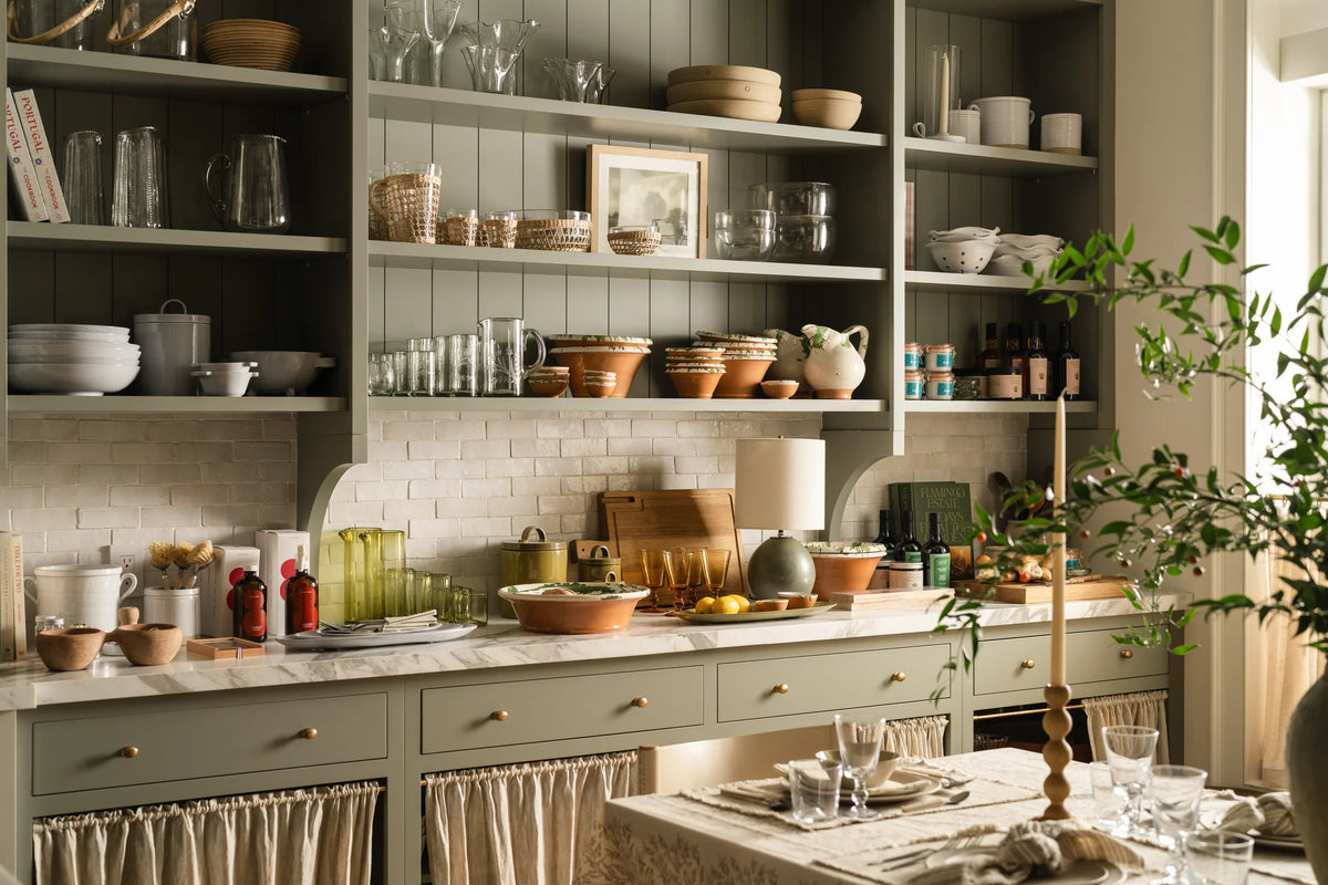 Sage green open shelving with glassware, terracotta and pottery above skirted lower cabinets in the Calimia Home showroom kitchen