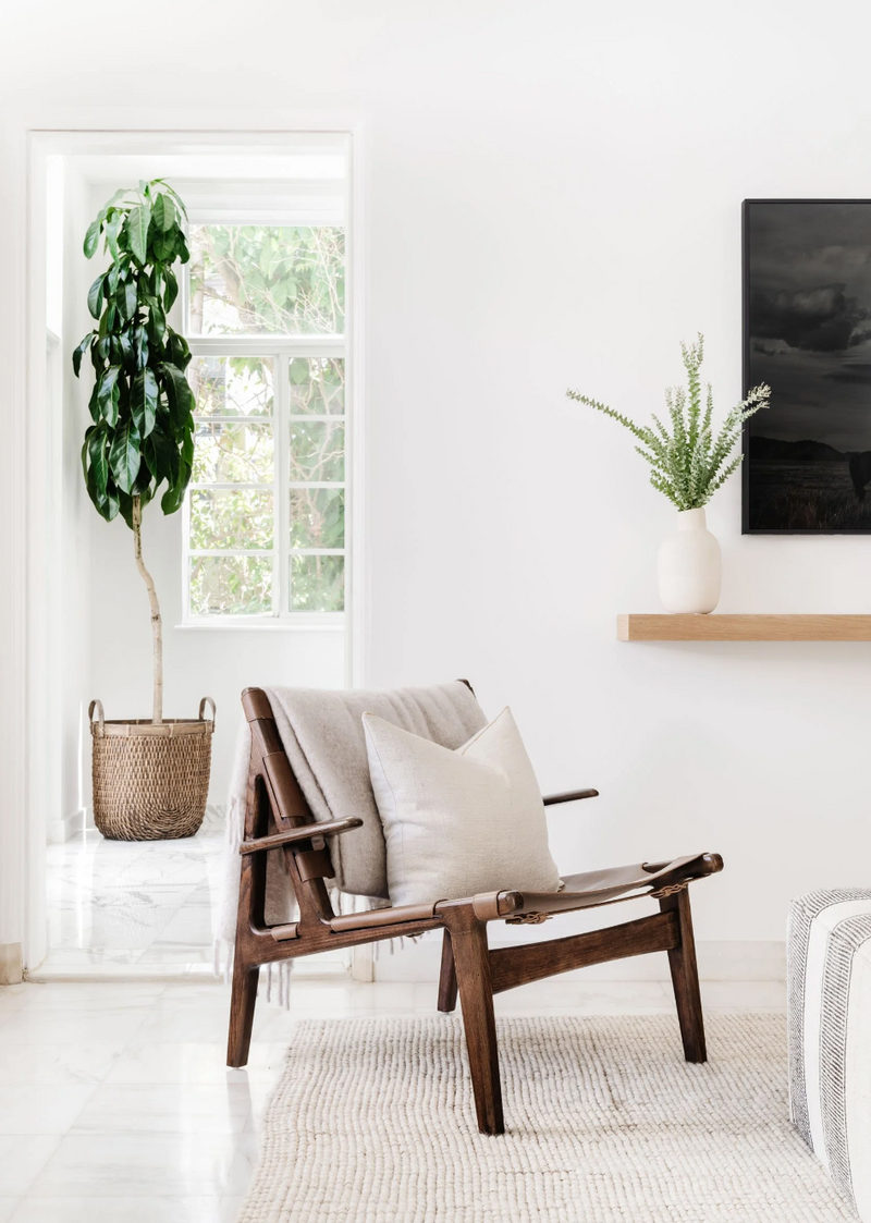 Walnut sling lounge chair with linen cushion beneath dark artwork and a potted tree