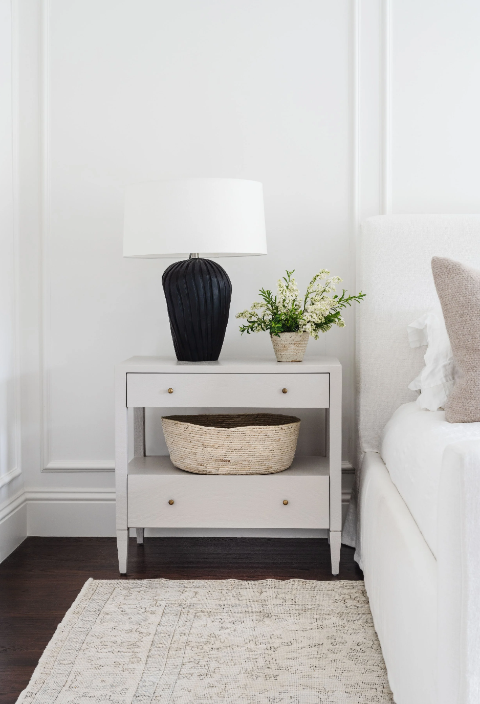 White nightstand with decorative items next to a bed in a bedroom setting.