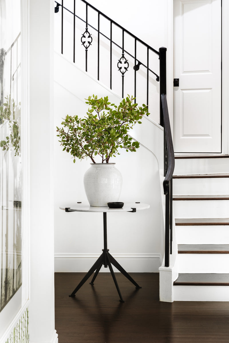 Marble-top pedestal table with greenery at the base of a wrought iron staircase