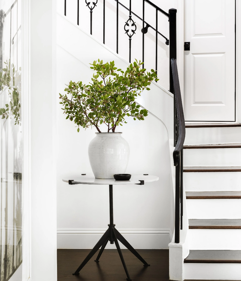White vase with green plant on a small round table against a staircase.