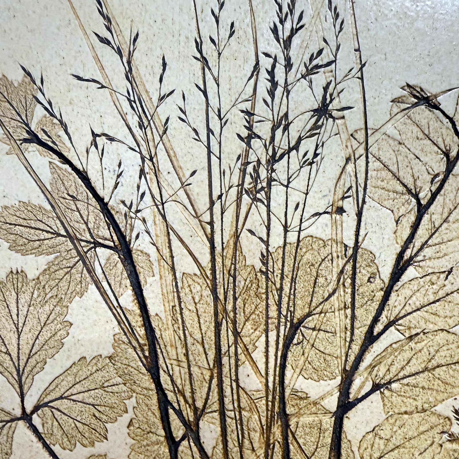 Close-up of a vintage ceramic botanical wall tile showing grass seed heads and leaf veins pressed in relief with an iron-toned wash