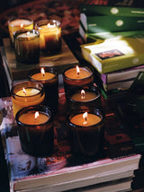 A cluster of lit Flamingo Estate Climbing Tuscan Rosemary Candles glows atop a stack of art books and magazines, with the brand's olive-green gift boxes layered behind.
