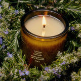 A brown glass Climbing Tuscan Rosemary Candle from Flamingo Estate burns brightly, cradled in a bed of fresh rosemary sprigs dotted with tiny pale-purple rosemary blossoms.