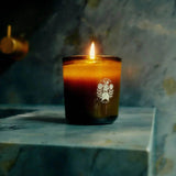A lit Flamingo Estate Douglas Fir & Ancient Vetiver Candle glows on a marble bath ledge, its flame reflecting brass fixtures behind in a smoky, woodsy still life.