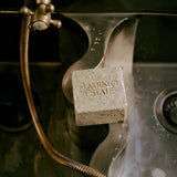 The Flamingo Estate Peppermint Bar Soap leans into a vintage metal sink basin, brass faucet and coiled hose dripping water beads down its grainy surface.