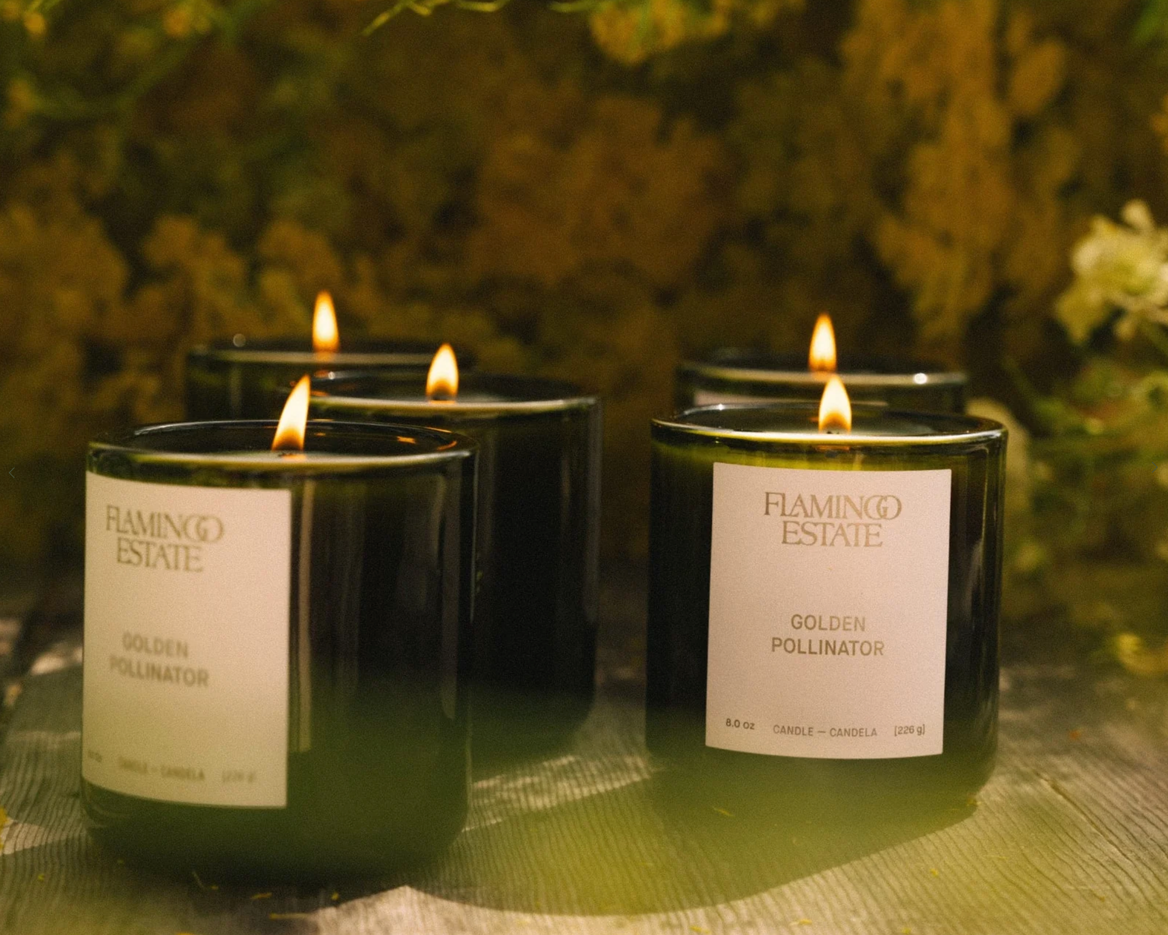 Flamingo Estate Golden Pollinator Candles burning on a wooden surface, showing flame and label detail
