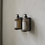 Calimia Home matte black wall-mounted bottle holder brackets holding two pump bottles on a neutral wall.