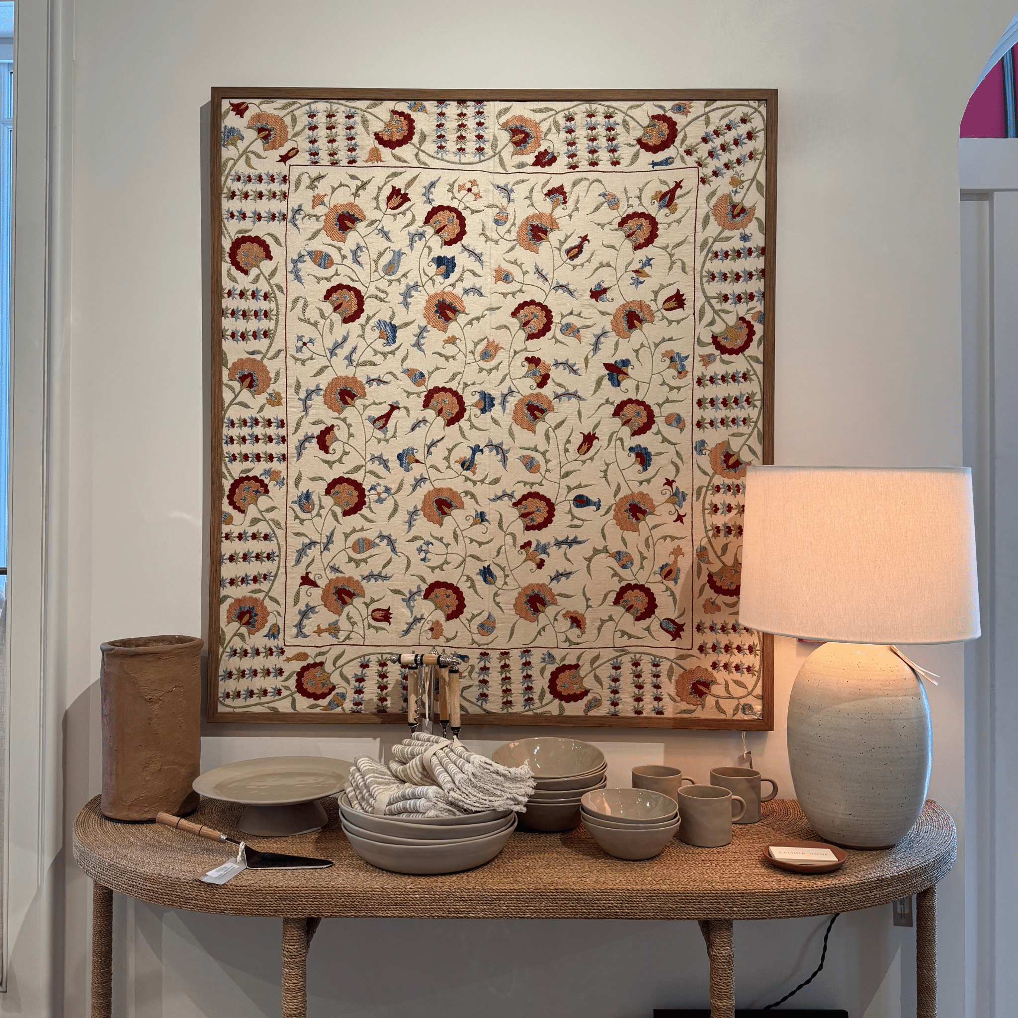 A console table with stacked dishes, cups, a striped towel, a ceramic lamp, and a brown paper bag sits beneath a Framed Vintage Suzani—vivid Central Asian embroidery—displayed on a white wall.