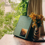 Fridays From The Garden cookbook props upright on a wooden table, framed by yellow wildflowers in a striped vase and trailing greenery beyond the window.
